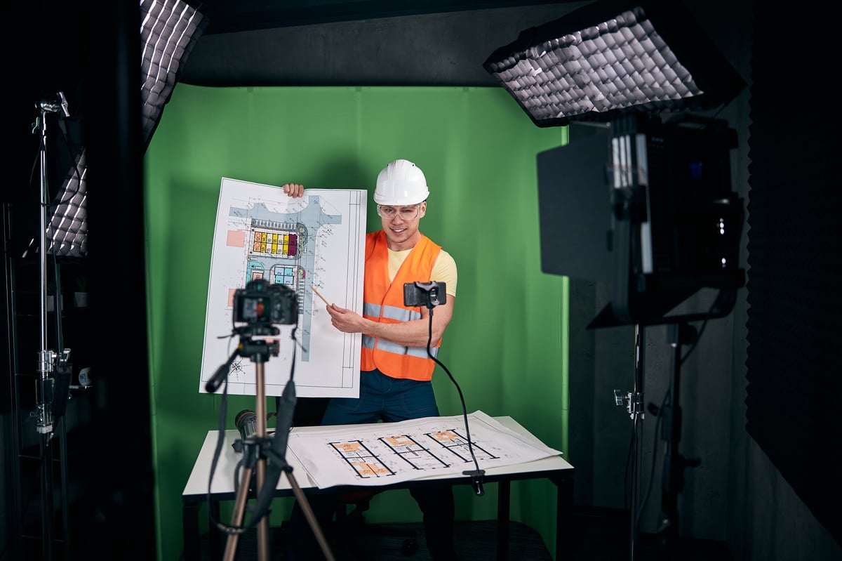 Handsome young man holding architectural plan and smiling while shooting video with professional camera
