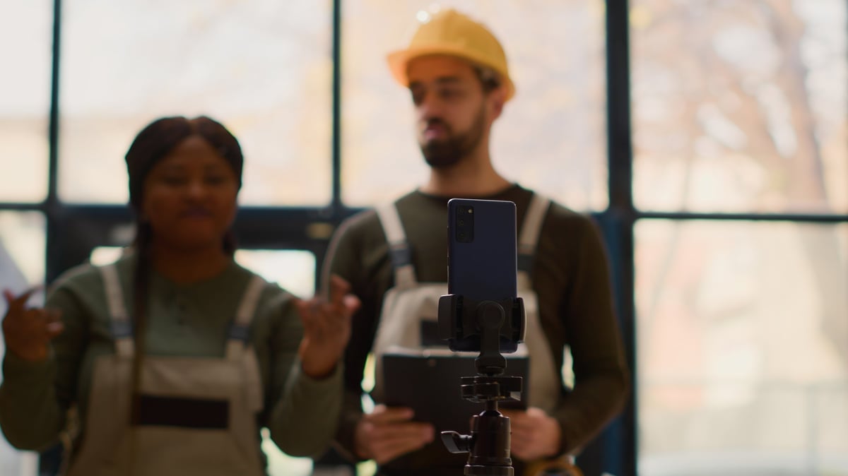 Focus shot on cellphone placed on tripod used for making training video for interns by warehouse colleagues in blurry background. Stuff members showing trainees how to seal packages